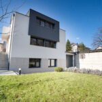 Kuća modernog dizajna pretežito bijele boje sa istaknutim crnim segmentom i velikim zelenim vrtom ispred