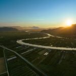 Široki zračni prikaz vijugave rijeke koja prolazi kroz zelenu poljoprivrednu dolinu u zalazak sunca. Rijeka stvara zmijoliki uzorak, dijeleći velika polja i plastenike, dok se planine u pozadini djelomično ocrtavaju u silueti na zalazećem suncu