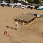 Fotografija drvenog beach bara na pješčanoj plaži, s parkiranim automobilima u pozadini.
