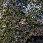 Pogled dronom iz visine na malu kuću s crvenim krovom usred guste šume i kamenitog krajolika