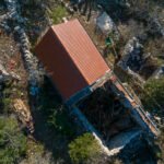 Izbliza snimka dronom kuće s crvenim krovom, okružene ruševinama i gustom vegetacijom u šumovitom kamenitom području.