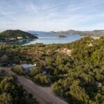 Prikaz obalnog brdovitog terena sa stablima i niskim grmljem. U prvom planu je zemljana cesta s malom zgradom sa strane. U pozadini se vidi mirna uvala s otočićima, a iza njih uzdižu se planine pod lagano oblačnim nebom