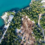 Pogled iz zraka na obalni krajolik s tirkiznim morem uz šumovito područje. Nekoliko zgrada nalazi se uz obalu, a staze vode kroz gustu vegetaciju. Područje ima mješavinu zelenila i dijelova gole zemlje s vidljivim stijenama