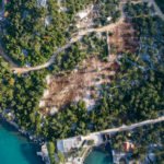 Zračni prikaz obalnog krajobraza s gustom zelenom vegetacijom, kroz koju prolazi zemljana cesta. U sredini je otvoreno, očišćeno područje golog tla. Na dnu, tirkizno more dodiruje kamenitu obalu s nekoliko manjih građevina uz obalu.