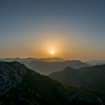 Miran planinski krajolik u vrijeme zalaska sunca, suncem koje zalazi iza niza maglovitih brda i planina. Nebo prelazi iz zlatnog sjaja na horizontu u tamnoplavu boju iznad, stvarajući mirnu atmosferu nad hrapavim terenom i šumovitim padinama.